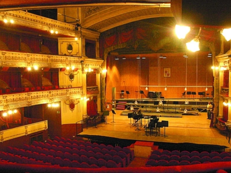 Interior of an ornate theater with a stage, red seats, and intricate ceiling details.
