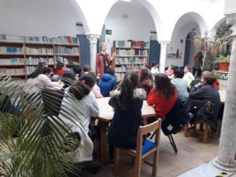 Escena de una biblioteca con arcos y estanterías llena de libros. Personas sentadas en mesas, algunas leyendo, otras conversando. Ambiente acogedor con plantas y decoración tradicional.