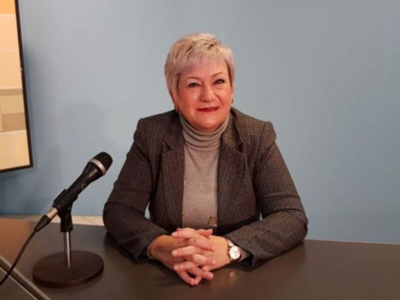 Una mujer con cabello corto y gris, vistiendo una chaqueta de cuadros marrones y un suéter beige, está sentada frente a una pantalla de ordenador y un micrófono. La pared al fondo es azul claro.