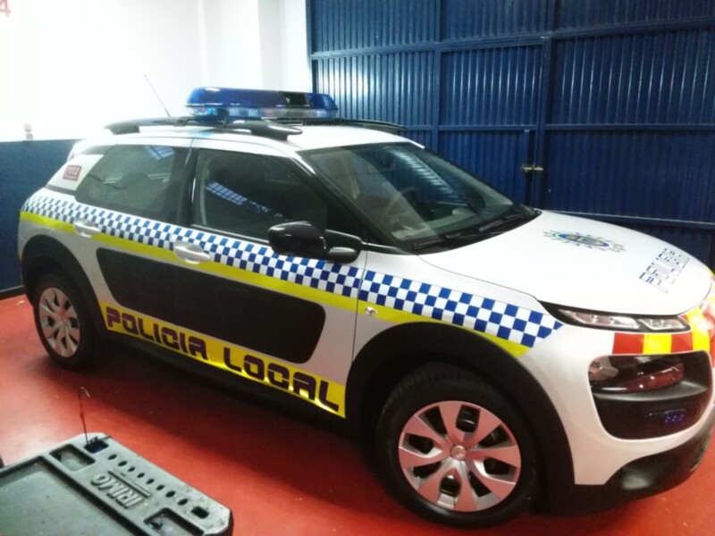 Un vehículo de policía estacionado en un hangar, con detalles de color azul y amarillo. La imagen muestra una vista frontal del vehículo, enfocándose en su diseño distintivo y el texto "POLICÍA LOCAL" en la parte delantera.