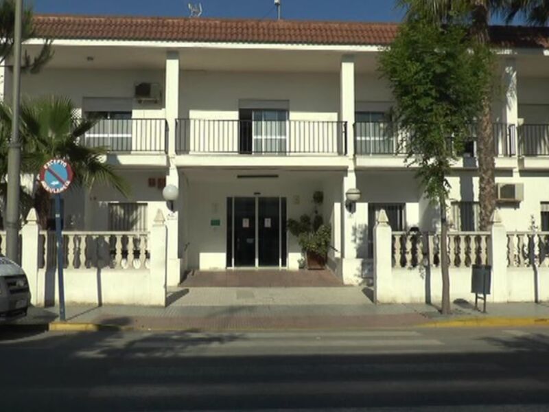 Fachada de un edificio blanco con balcones y ventanas, junto a una calle con árboles y un coche estacionado.