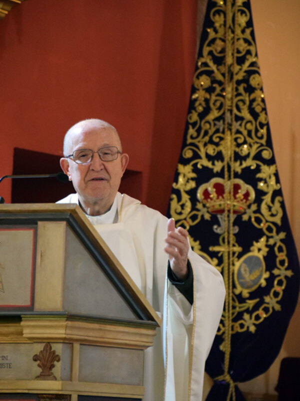 Un sacerdote en una iglesia, hablando en un púlpito con un fondo rojo y una bandera con un escudo de armas en la parte superior.