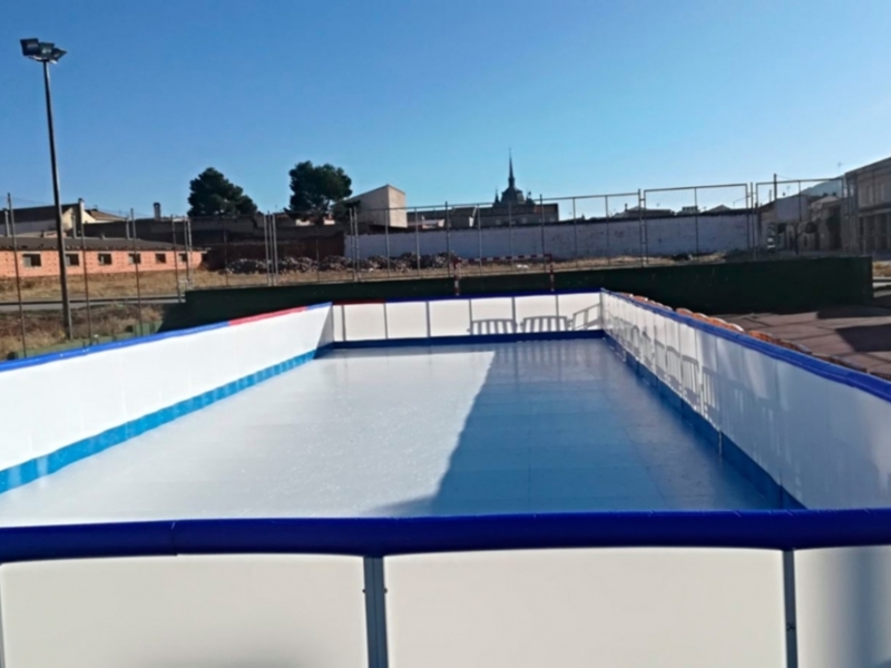 Una piscina rectangular con líneas azules y blancas, rodeada por un perímetro de metal. La piscina está situada en una zona abierta con edificios y árboles al fondo. La vista es clara y soleada, con un cielo azul claro.