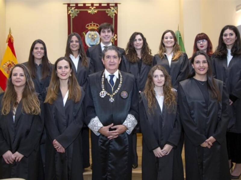 Equipo de profesionales en traje negro, posando frente a una bandera española y un escudo heráldico. La imagen sugiere un entorno formal, posiblemente en un entorno institucional o profesional.