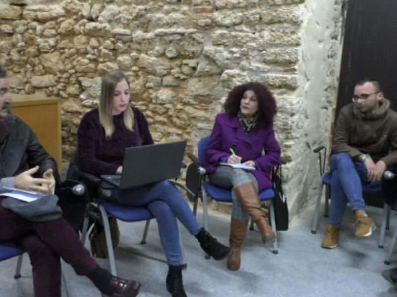 Una reunión de trabajo en un entorno de oficina con una pared de piedra y sillas. Los participantes están sentados, algunos con portátiles abiertos, mientras otros tienen documentos en sus manos. La imagen transmite un ambiente de colaboración y trabajo en equipo.