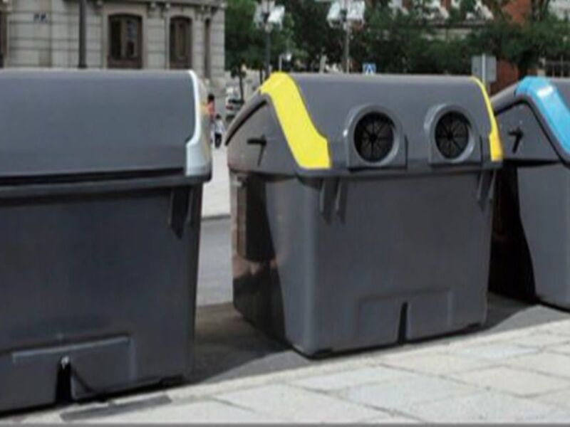 Tres contenedores de reciclaje en una calle con edificios y árboles al fondo.