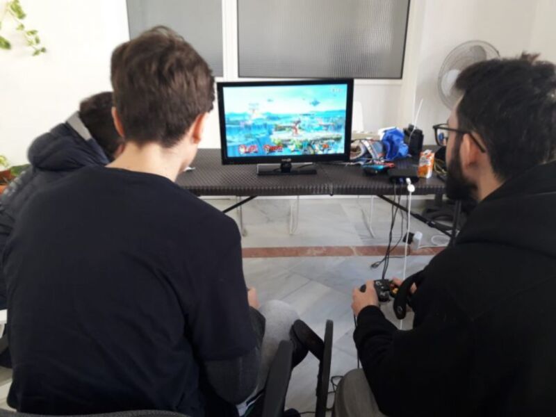 Dos personas están sentadas frente a un monitor de videojuego, jugando una partida. La escena se centra en la interacción y el entretenimiento de los jugadores, con una tableta y un ventilador en la mesa.