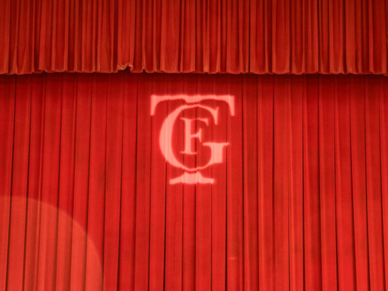 Cortina roja con logo CFG en el centro, representando un escenario teatral o presentación formal.