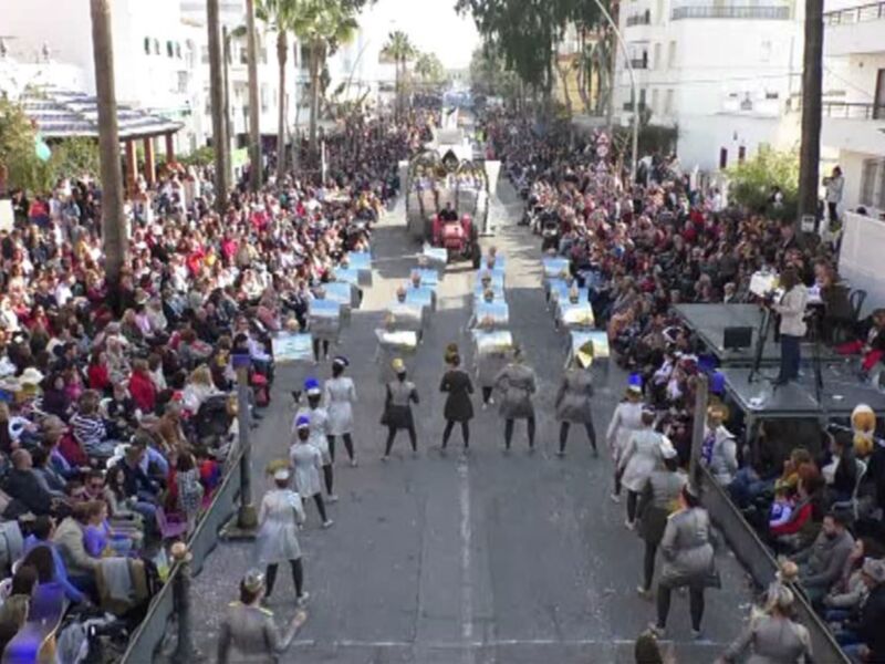 Desfile de carros alegóricos y danza en una calle llena de espectadores durante un evento festivo.