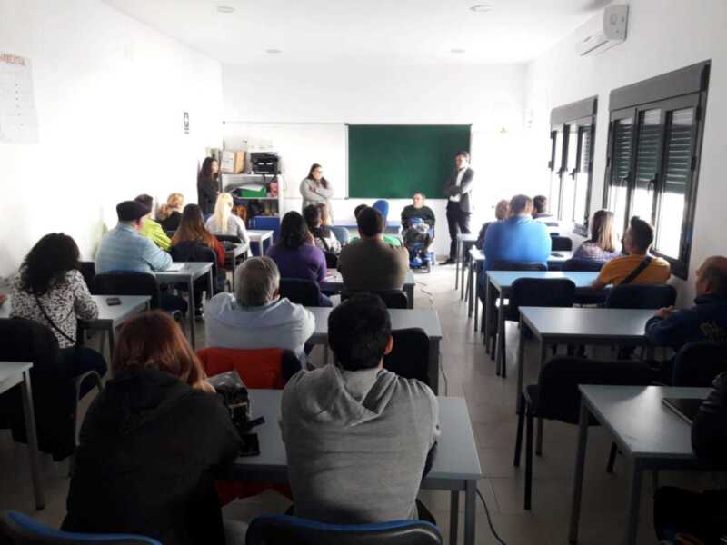 Clase de idioma en un aula con estudiantes sentados en mesas. Un profesor está enseñando frente a una pizarra verde y un tablero blanco con un calendario. La sala tiene ventanas laterales, sillas rojas y azules, y una mesa con un portátil.