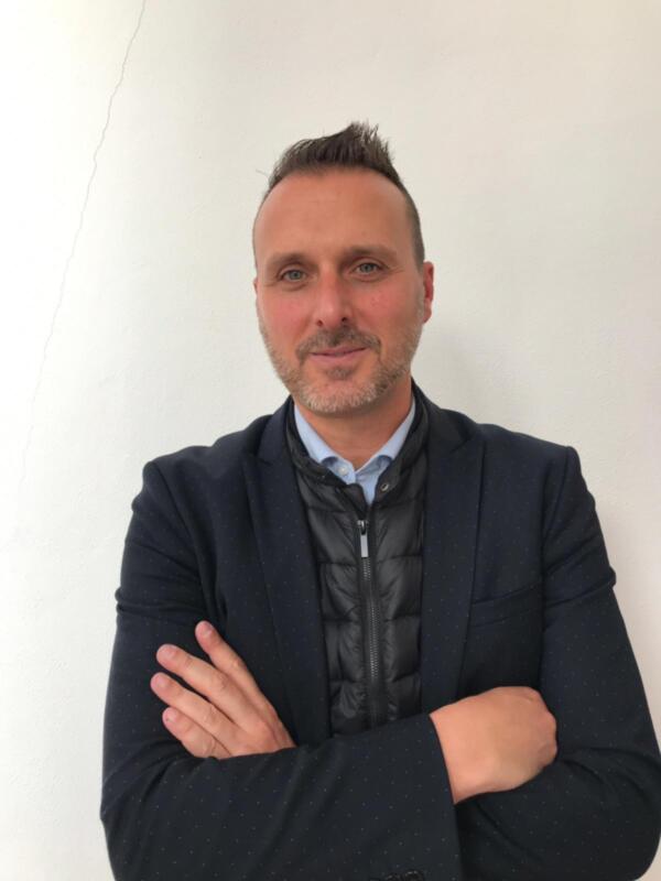 Un hombre con cabello corto y una barba gris, posando con los brazos cruzados en frente de un fondo blanco. Se viste con una chaqueta azul y una camisa interior de color claro, mirando directamente a la cámara.