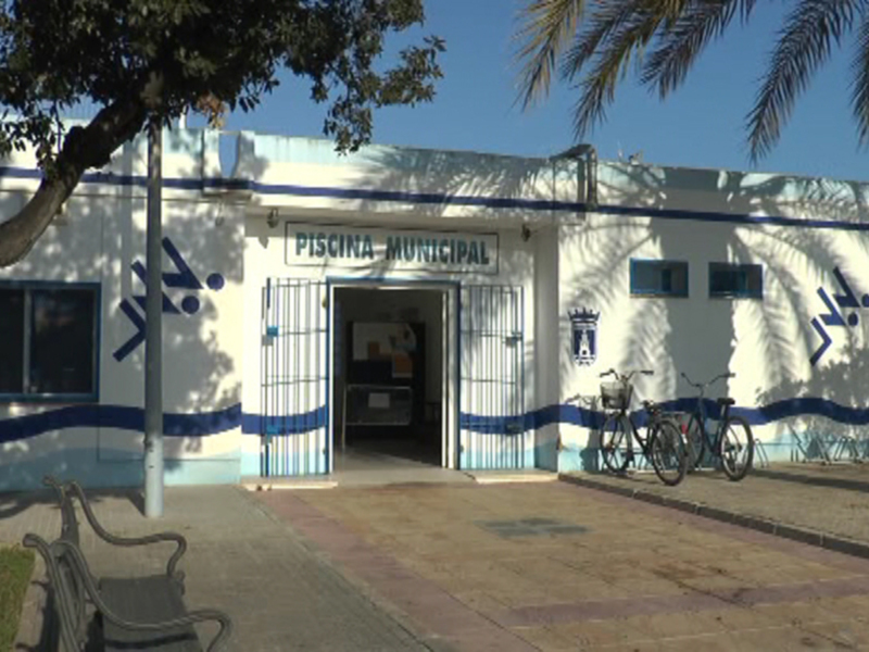 Piscina Municipal con bicicletas y árboles frente a un edificio blanco bajo un cielo azul claro.