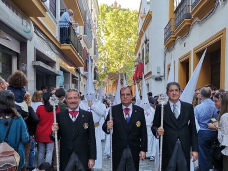 Procesión religiosa con participantes en trajes formales y túnicas blancas, celebrada en una calle urbana.