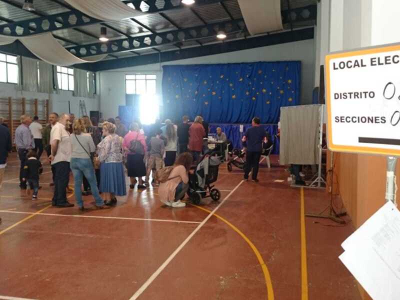 Votación en un local electoral, con personas esperando en fila y un cartel indicando las secciones de votación.