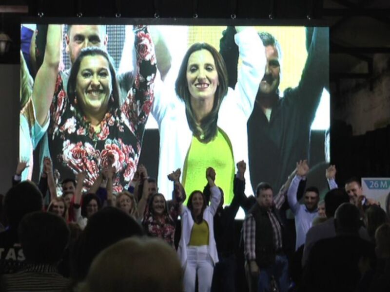 Celebración en un evento público con personas levantando las manos y una pantalla grande en el fondo mostrando a tres personas felices.