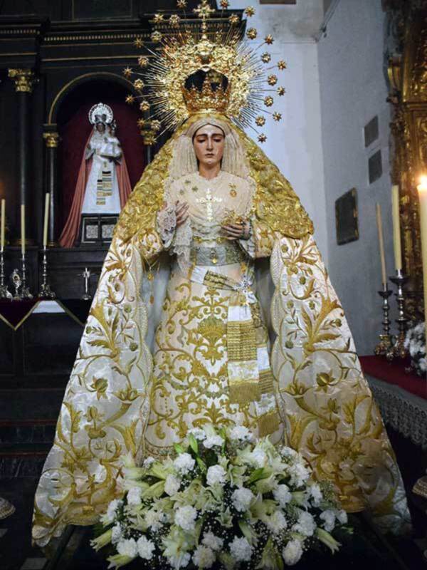 Imaginación de la Virgen María en una iglesia, vestida con una magnífica capa dorada y blanca, rodeada de flores y con un altar en el fondo.