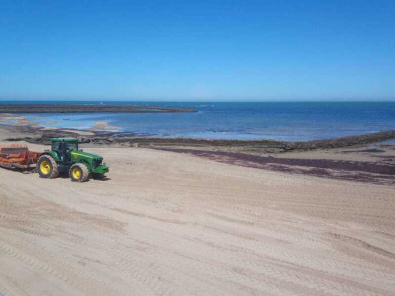 Un tractor verde y amarillo está trabajando en una playa arenosa, con el mar y la costa al fondo. El cielo es azul claro.