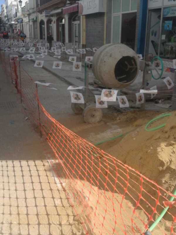 Se observa una calle con un muro de hormigón en construcción. Al fondo, hay edificios comerciales y una señalética publicitaria de "Mundo Móvil". La calle está bloqueada con un muro de hormigón y una red de seguridad.