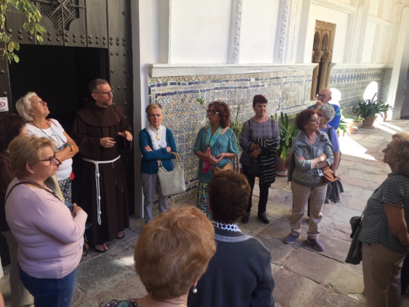 Una imagen de un grupo de personas en una visita guiada, posiblemente en un museo o edificio histórico. La gente está concentrada y escuchando a un guía, mientras miran hacia la entrada principal. La decoración del edificio muestra azulejos y detalles arquitectónicos tradicionales. La atmósfera es educativa e interesante, con una mezcla de personas de diferentes edades y estilos.