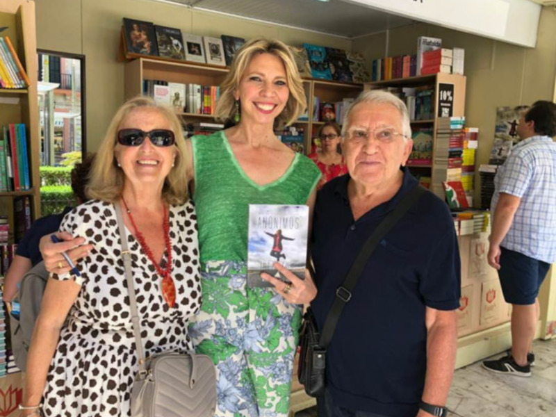 Tres personas sonrientes en una librería, una sostiene el libro 'Anónimos'.