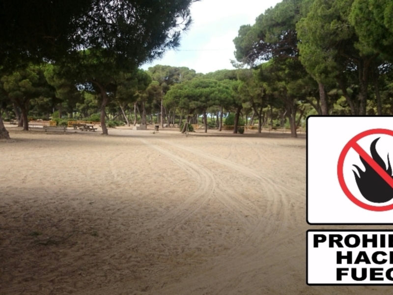En un tranquilo parque con árboles altos y una zona de arena, se observa un letrero que prohíbe hacer fuego.