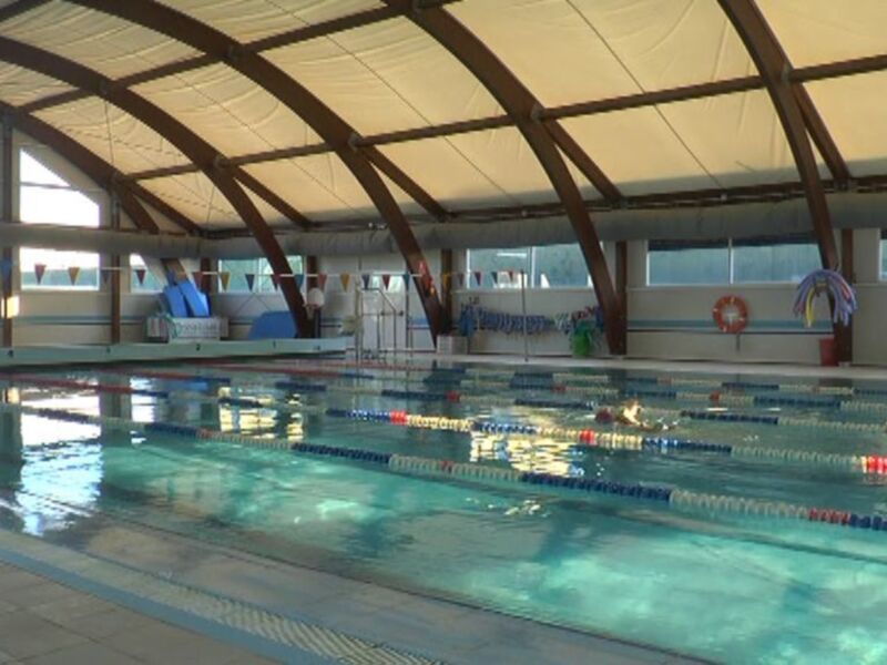 Piscina interior con natación y estanques de hidromasaje. Paredes blancas, techo curvo con iluminación natural y reflejos en el agua.