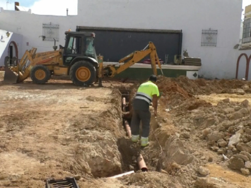 Trabajadores excavando en una zona de construcción, con una excavadora amarilla en el fondo y una casa blanca a la izquierda.