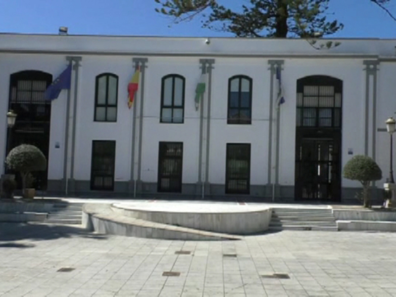 Fachada de un edificio blanco con ventanas y puertas negras, banderas en la fachada. Suelo de piedra y árboles al lado.