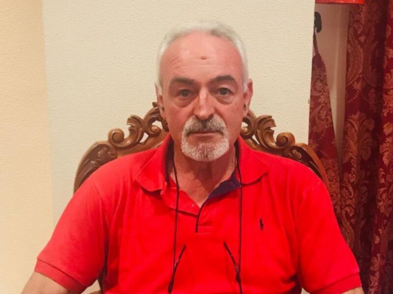 Un hombre con barba gris y una camisa roja está sentado en un sillón de madera. La pared detrás de él es beige y hay una cortina roja con un patrón floral.