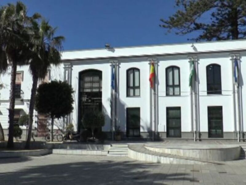 Un edificio blanco con ventanas y dos banderas en el exterior. En la entrada, hay una escalera de piedra. Al fondo, se pueden ver árboles y un cielo azul claro.