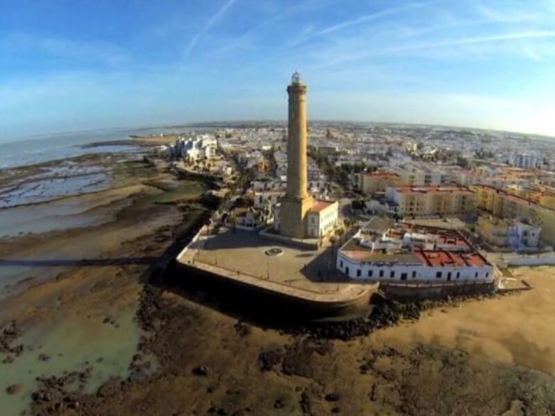 Una vista aérea de la ciudad de Cádiz, España. Se puede observar la Mezquita-Catedral y el faro de Cádiz, ubicados en una bahía. La vista incluye la costa y el horizonte despejado, con un cielo azul claro.