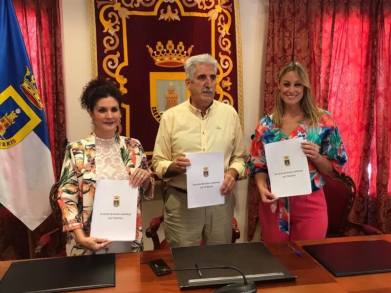 Una imagen de tres personas en una oficina, con un hombre y dos mujeres. La mujer izquierda sostiene un documento con el escudo de armas, la mujer derecha también sostiene un documento similar. La oficina tiene una bandera y cortinas rojas en el fondo, con un escritorio de madera.