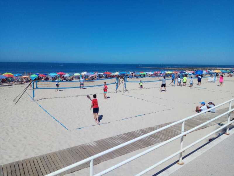 Una imagen de una playa con varios juegos de voleibol en acción. La arena es clara y el cielo está despejado con un azul profundo. Hay varios jugadores en diferentes posiciones, algunos están en el aire intentando alcanzar la pelota. También se pueden ver varios paraguas de colores y personas disfrutando del día en la playa. La imagen captura el entusiasmo de los jugadores y la relajación de los espectadores.