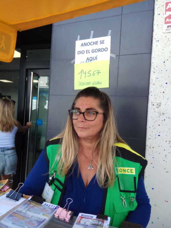 Una mujer con una chaqueta verde y azul está frente a un edificio con un cartel de apoyo al gordo. La imagen muestra una persona en una tienda o centro comercial, posiblemente esperando resultados.