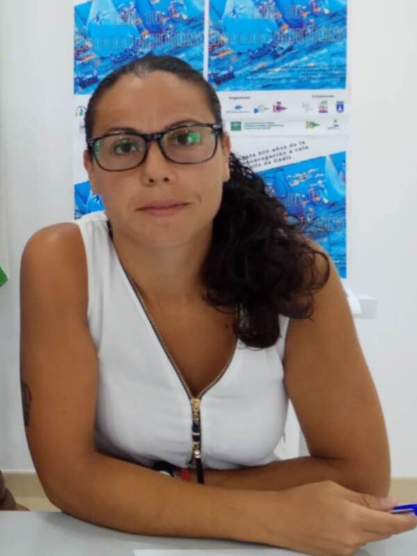 Mujer con gafas y blusa blanca sentada en una mesa con carteles de fondo.