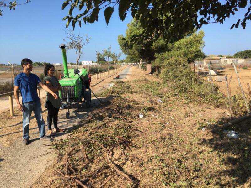 Dos personas caminan junto a un tractor verde en medio de una zona despejada, con árboles y vegetación alrededor. El cielo es azul claro.