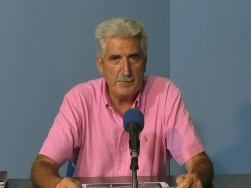 Un hombre con cabello blanco y bigote, vestido con una camiseta rosa, se sienta frente a un micrófono en un fondo azul. En la mesa delante de él hay un libro y gafas de sol. La imagen sugiere una situación formal o informativa, posiblemente un discurso o entrevista.