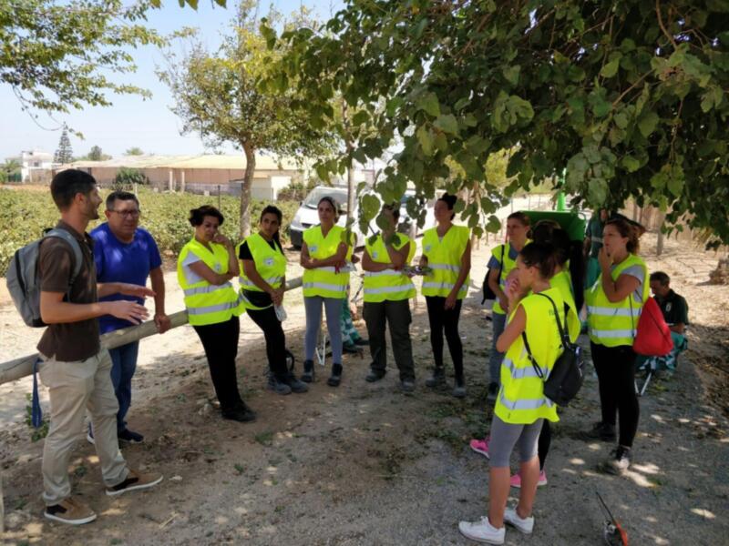 Un grupo de personas en chalecos reflectantes están reunidas alrededor de un hombre que parece estar explicando algo. La escena se desarrolla en un entorno natural con árboles y vegetación al fondo. La gente parece estar atenta a la explicación, posiblemente en un contexto educativo o de capacitación.