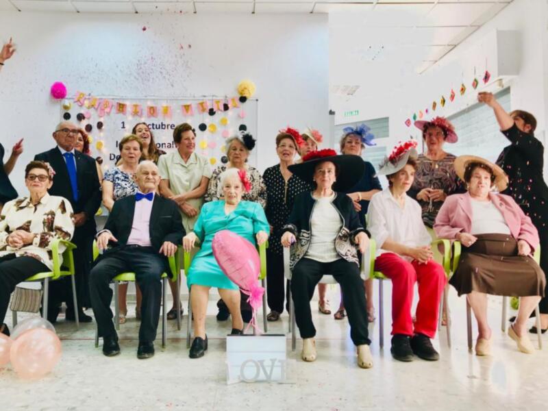 Una celebración de cumpleaños en un centro de jubilados, con personas vestidas con trajes elegantes y sombreros coloridos, rodeadas de globos y confeti. La fecha es el 1 de octubre.