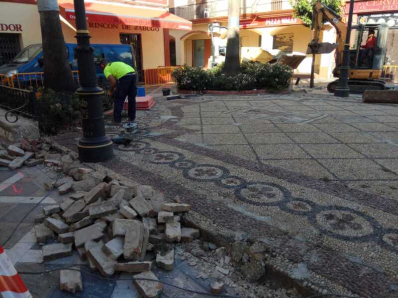 Trabajadores en un área de construcción, con una excavadora y un camión de volteo. La imagen muestra una calle con adoquines rotos y una pila de ladrillos. En el fondo, se pueden ver edificios con toldos rojos y una escena de trabajo en la ciudad.