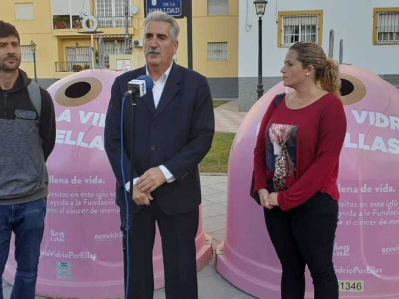 En un entorno urbano, tres personas se encuentran frente a dos grandes contenedores de vidrio rosa. El ambiente es relajado, con un hombre vestido con una chaqueta negra y vaqueros, una mujer en una camiseta roja con estampado floral y un hombre vestido con traje negro. El fondo muestra edificios de viviendas y una calle con vegetación, todo bajo un cielo despejado.
