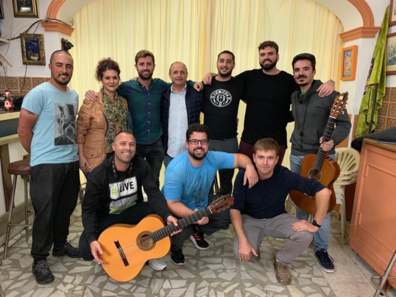Grupo de personas reunidos en una habitación, algunos con instrumentos musicales, sonrientes y felices.
