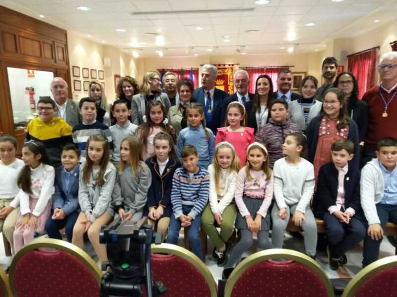 Una escena de una reunión en un salón con niños y adultos sentados en filas. La imagen muestra a varias personas, incluyendo niños de diferentes edades y adultos, sentados en filas. La sala tiene una decoración formal con cortinas rojas y un fondo que parece ser una pared de madera. La iluminación es buena, lo que permite ver a todos los individuos claramente. En la parte inferior de la imagen, se puede ver una cámara de video en el centro del salón.