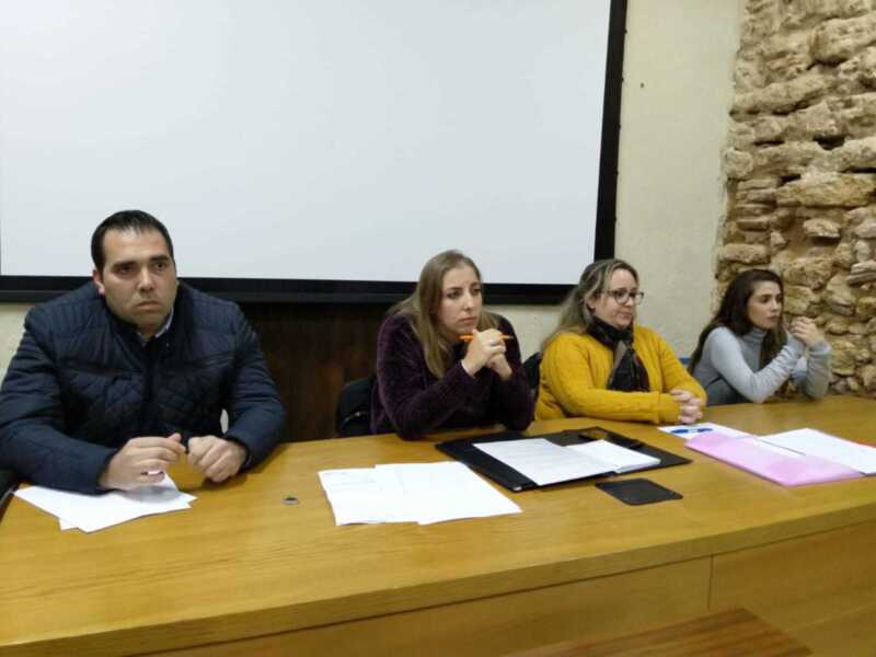 Una imagen de cuatro personas sentadas en una mesa, con un hombre y tres mujeres. La mujer del centro sostiene un marcador de color naranja, mientras que las otras dos mujeres tienen sus manos en el aire. La mesa tiene documentos y un marcador de color naranja. En el fondo, se puede ver una proyección en la pared y una pared de piedra.