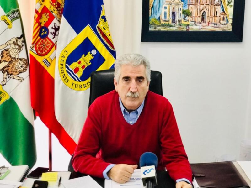 Persona en traje rojo sentada en una oficina con banderas y un retrato de edificio en el fondo.