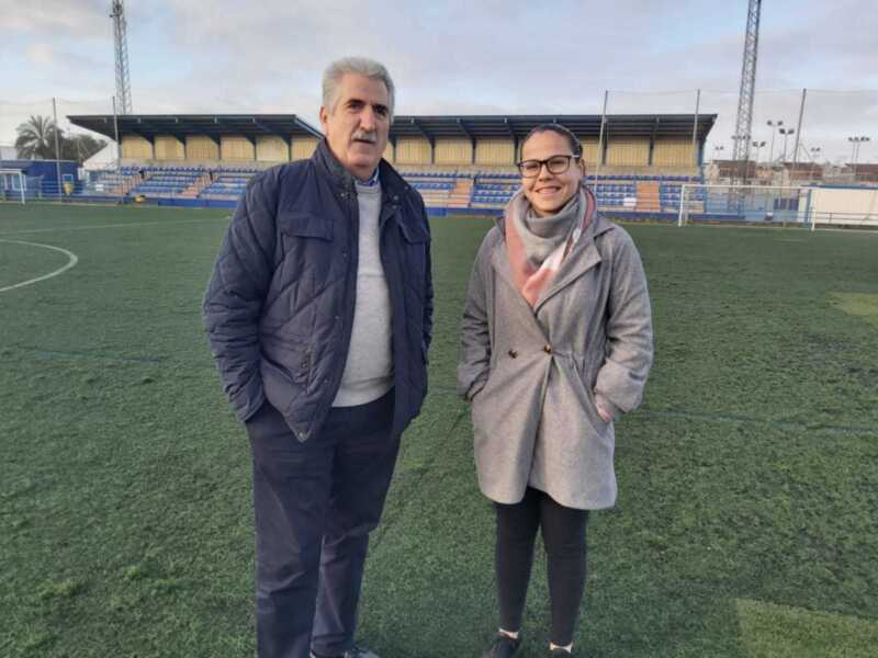 Dos personas posan en un campo de fútbol con una tribuna vacía al fondo. La imagen captura un momento casual en un entorno deportivo.