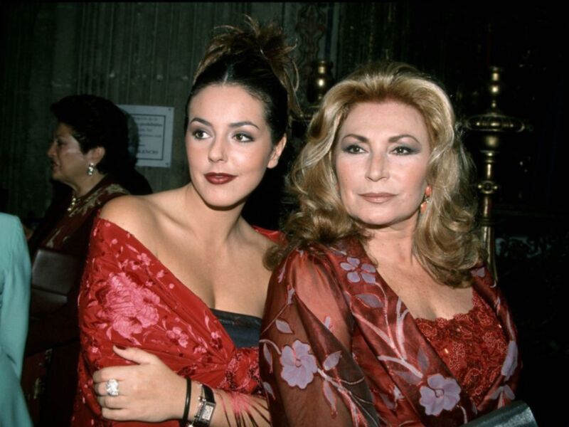 Dos mujeres jóvenes y una anciana posan juntas en un evento formal. La mujer joven se viste con un vestido rojo floral, mientras que la anciana lleva una prenda similar. Ambas tienen vestidos de gala y son posibles actrices o celebridades. La escena transmite elegancia y amistad entre las tres personas.