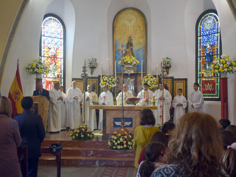 Celebración religiosa en una iglesia con ofrendas florales y ofrendas en el altar, con personas vestidas con hábitos religiosos y oficiales en el fondo.