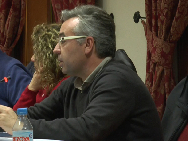 Un hombre con gafas y una camisa negra está sentado en un escritorio, rodeado de documentos y una botella de agua. La imagen parece ser de una reunión o conferencia formal, con cortinas rojas en el fondo.