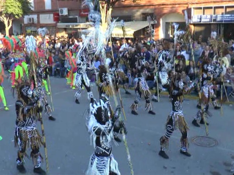 Desfile de danza tradicional con vestimentas coloridas y máscaras en una calle animada.